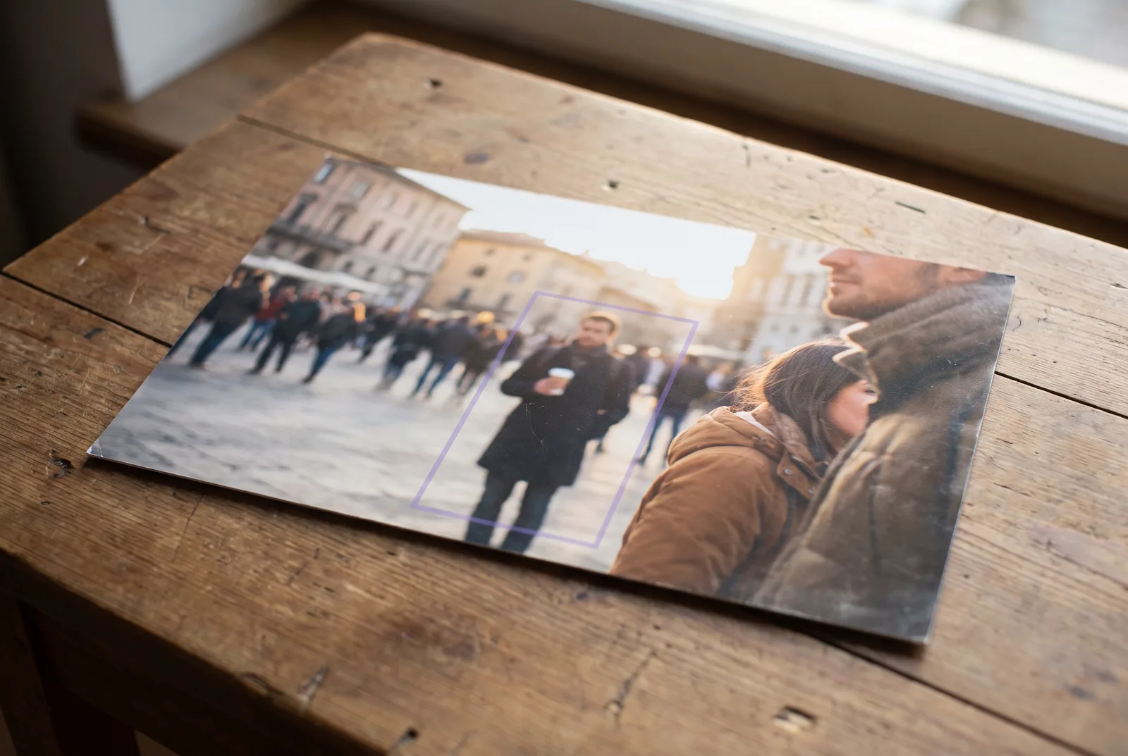 A printed holiday photo of a couple in a sunny plaza, faces cropped out of frame. In the blurred crowd behind them one passer-by holding a coffee is outlined by a faint violet detection rectangle.