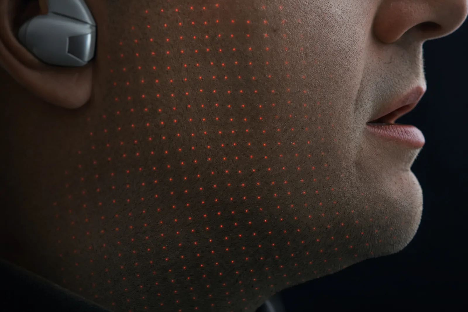Extreme close crop of an adult's cheek and jaw in profile, with a faint grid of structured-light sensing dots projected across the skin and a wearable earpiece at the edge of frame