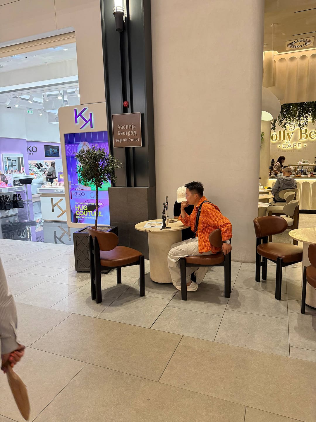 Inside a Belgrade mall, a person at a café table filming with a phone on a small tripod.