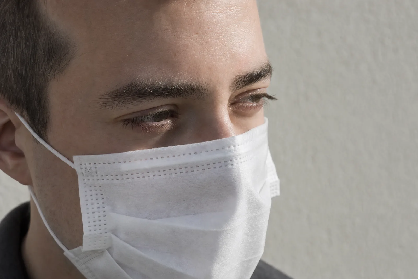 A close daylight portrait of a man in a plain white disposable mask covering nose, mouth and chin. The mask is soft-focused while the exposed strip of eyes, brows and upper cheeks is sharp and brightly lit.