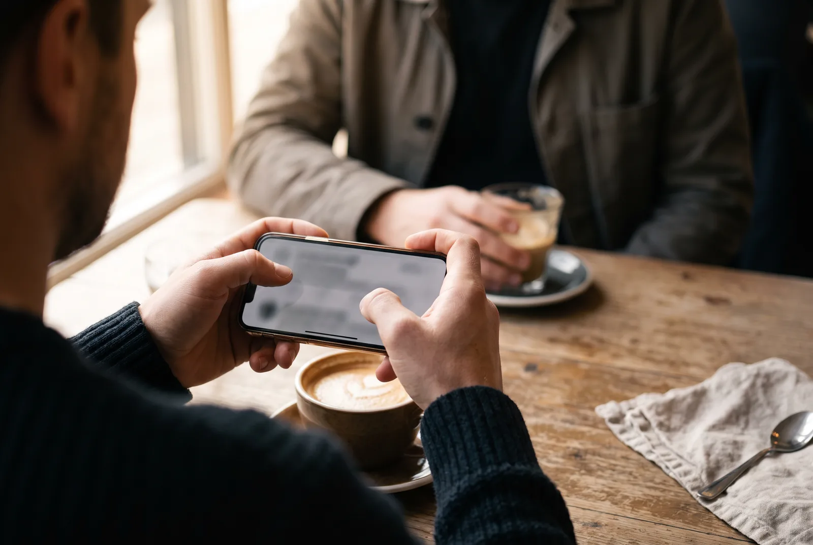 Over the shoulder of an adult typing a message on a phone at a cafe table, with a second person out of focus across the table.